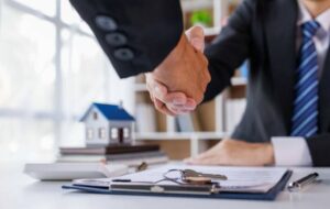 Two business professionals shaking hands over a desk with paperwork and a model house in the background, symbolizing an agreement related to a change of use in property. The scene suggests a formal real estate or planning decision.